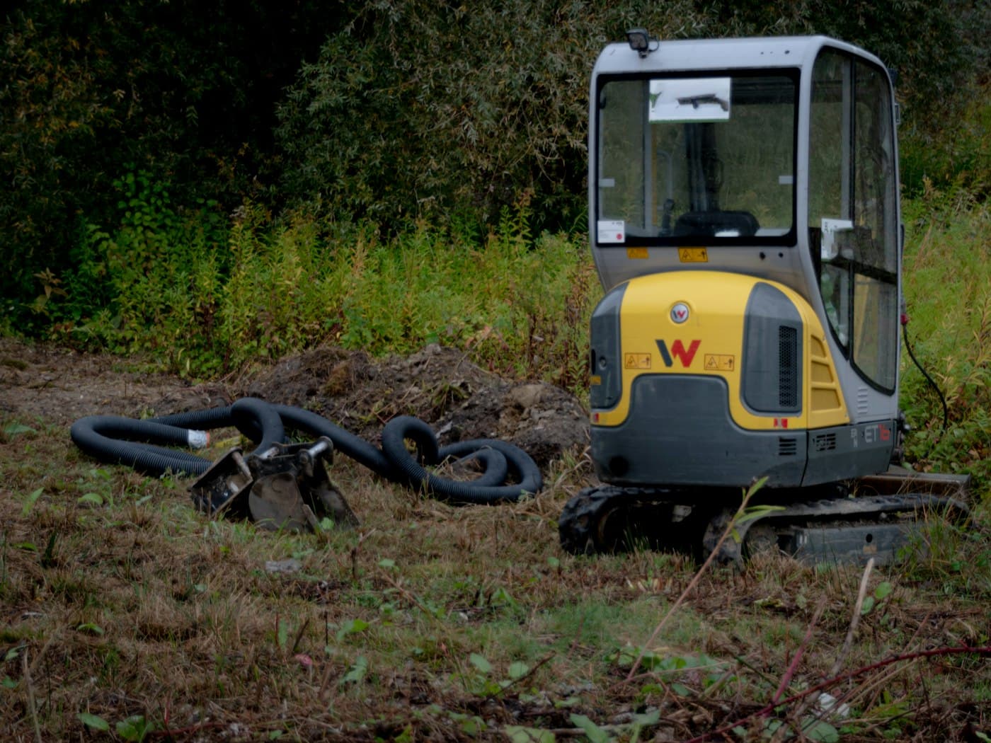A mini excavator parked at the edge of a work area with hoses coiled beside it.