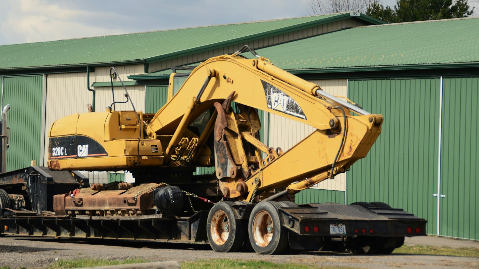 An excavator chained down on a lowboy trailer outside a metal shop building.